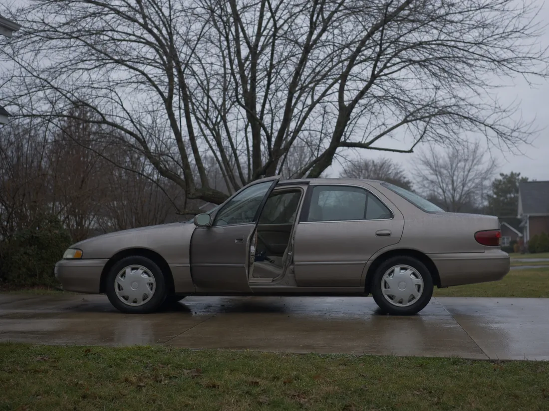 An older tan sedan standing on a wet driveway under a bare tree