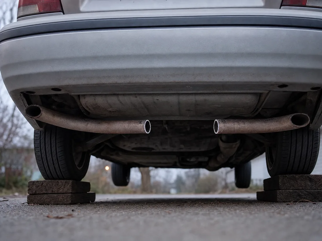 The rear underside of an older silver sedan raised on blocks, two freshly cut exhaust pipe ends facing the camera with a clean gap between them where the catalytic converter used to sit