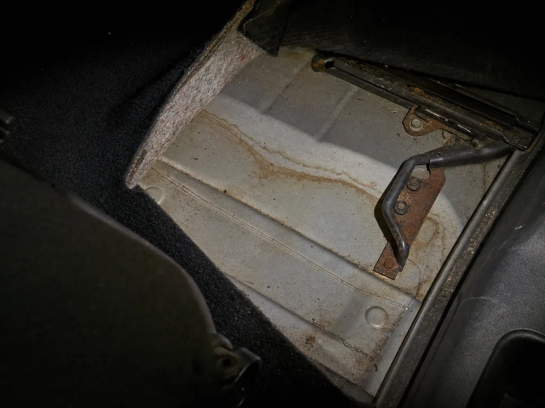 A car footwell with the carpet peeled back to bare metal, a dried silt tide-line across the floor pan and rust on the seat rail under a work torch
