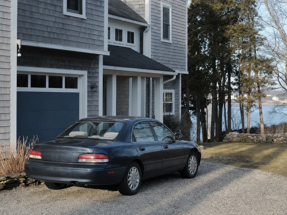 Older sedan beside a gray-shingled coastal home in Groton Connecticut with a glimpse of harbor water past the trees - selling a car in Groton