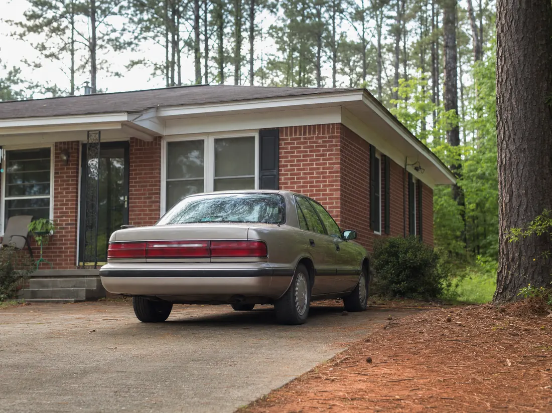 Older sedan at a modest brick home near Columbus with pines and red-clay driveway edges - selling a car in Columbus GA