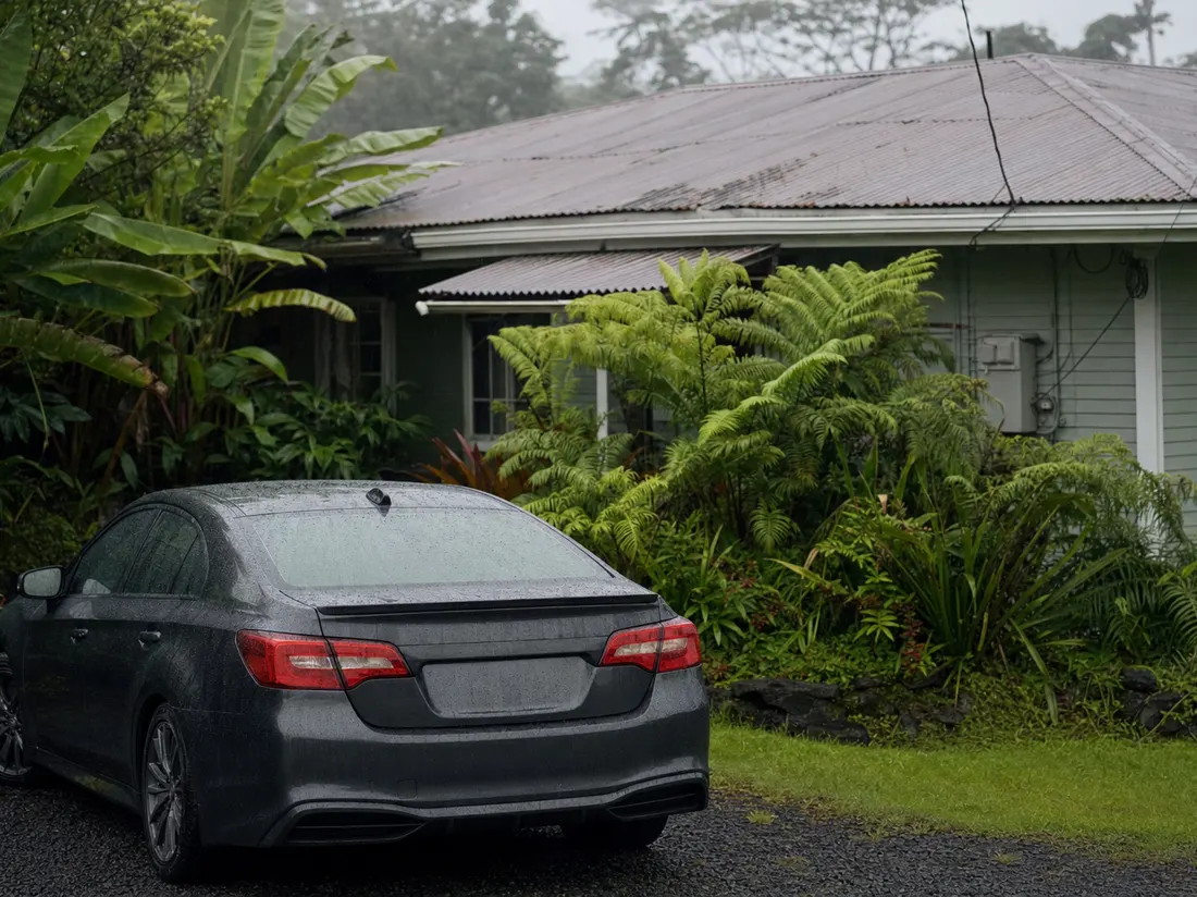 Sedan beaded with rain beside a tin-roofed Hilo Hawaii house surrounded by lush ferns and banana leaves - selling a car in Hilo