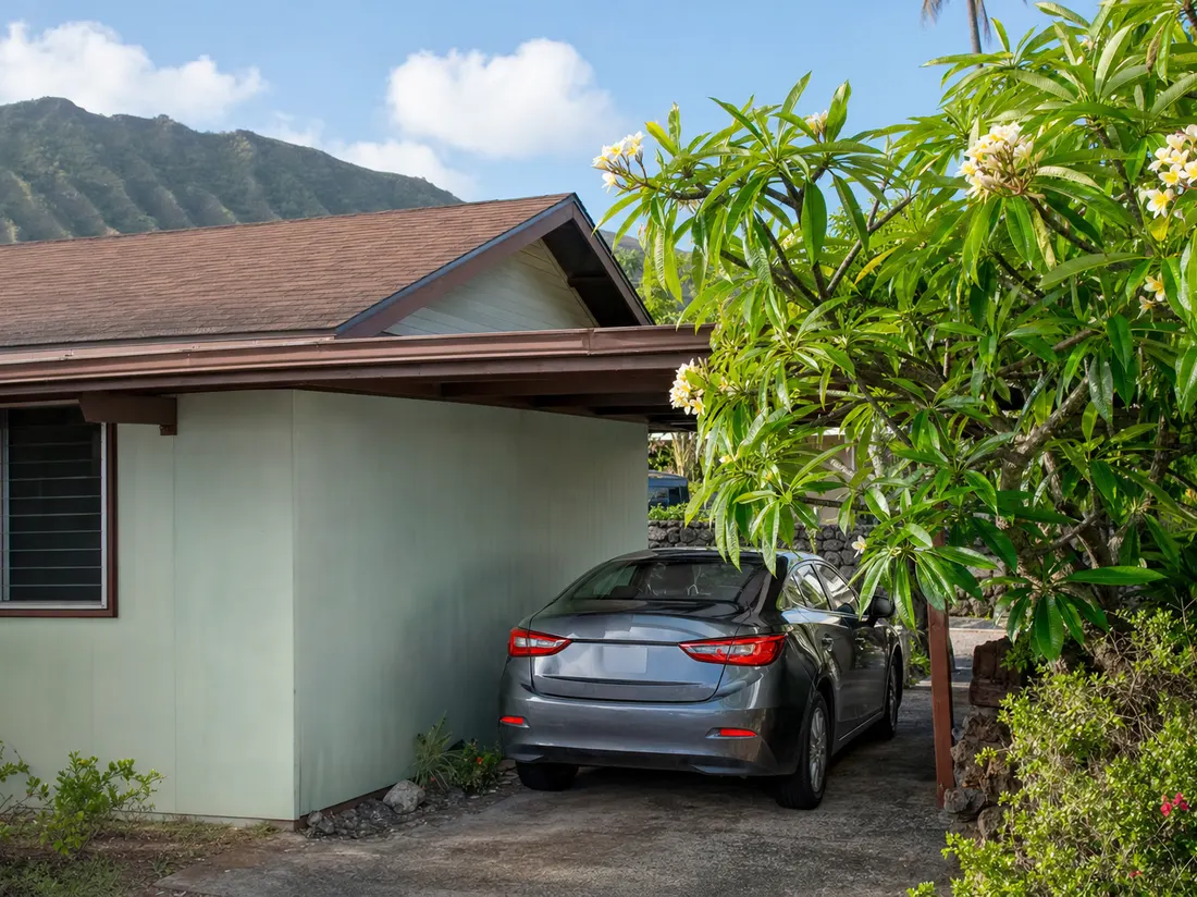 Compact sedan in the tight carport of a single-wall Honolulu Hawaii home with plumeria blossoms and a green ridgeline behind - selling a car in Honolulu