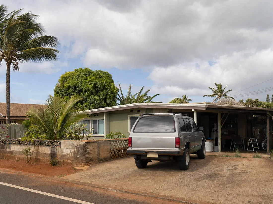Older SUV in the driveway of a modest Kahului Maui home with palms swaying and red-dirt shoulders on the street - selling a car in Kahului