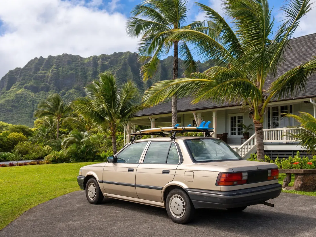 Small older sedan with surf racks parked at a plantation-style Hawaiian home with palms and volcanic ridge behind, bright trade-wind day - selling a car in Hawaii