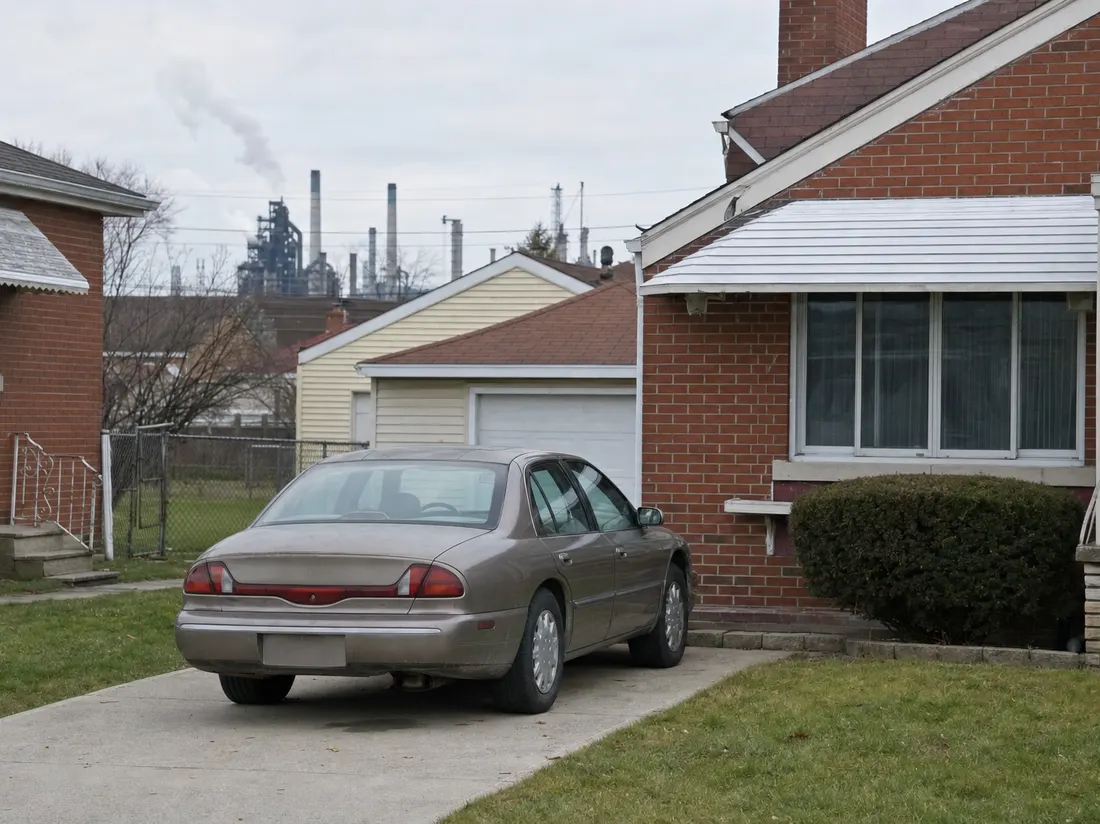 Older sedan in the driveway of a modest brick home in Gary Indiana with steel-mill stacks faint on the horizon past the rooftops - selling a car in Gary