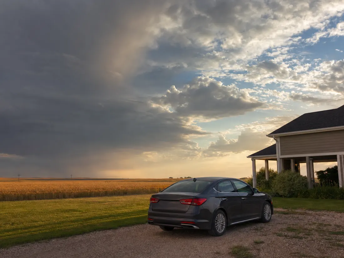 Sedan parked by a Kansas home under a huge dramatic prairie storm sky clearing to sun, wheat field behind - selling a car in Kansas
