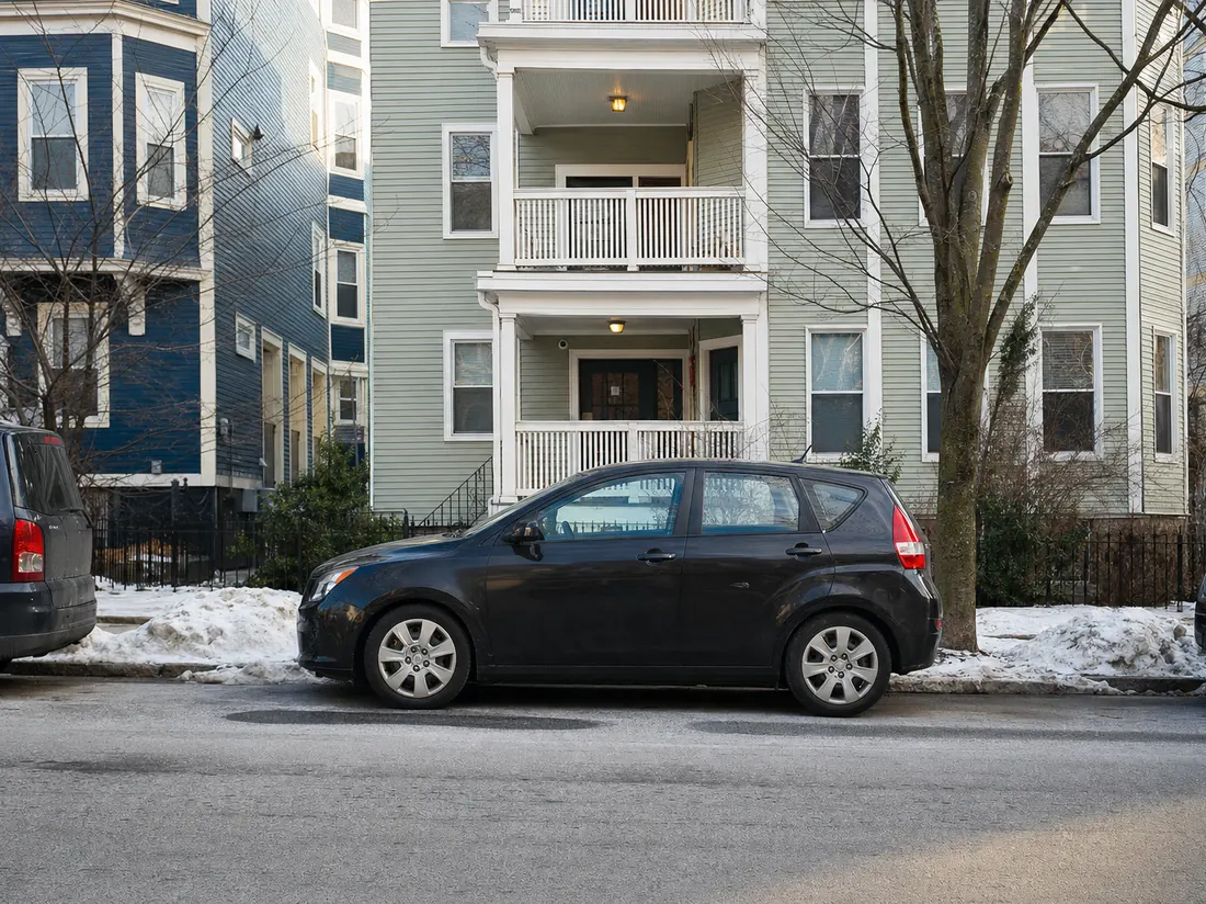 Compact car parallel-parked on a Boston triple-decker street with snowbank remnants at the curb - selling a car in Boston