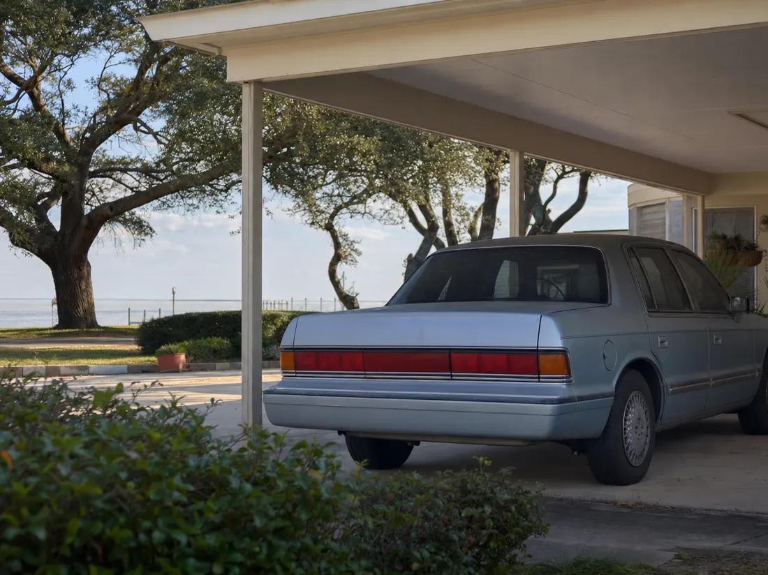 Older sedan under a carport at a low Biloxi Mississippi home with wind-shaped live oaks and gulf haze beyond - selling a car in Biloxi