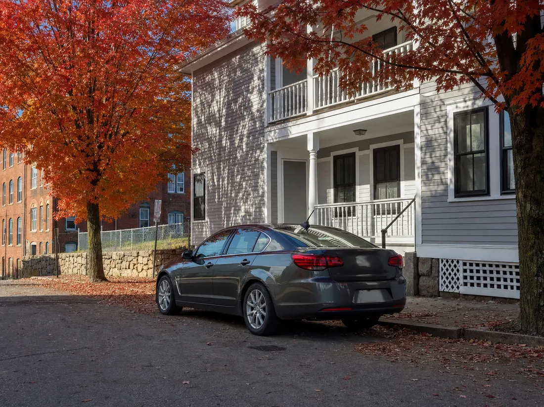 Sedan parked by a mill-town triple-decker in Manchester New Hampshire with fall maples reddening the block - selling a car in Manchester
