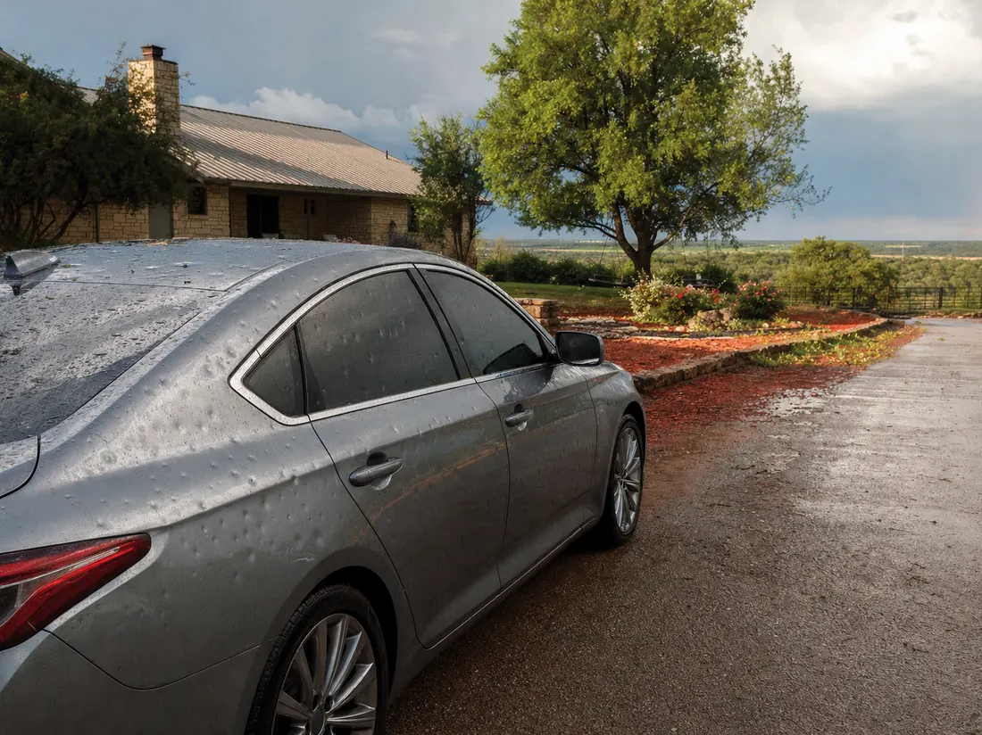 Hail-dimpled sedan at an Oklahoma City ranch home, red-dirt flowerbeds, storm clearing behind - selling a car in Oklahoma City