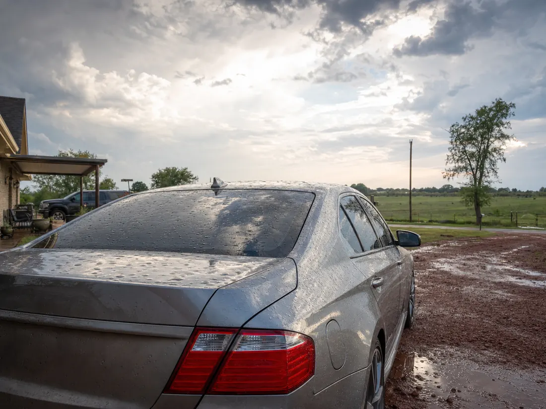 Sedan with small hail dents parked at an Oklahoma home under a big clearing storm sky, red-dirt driveway - selling a car in Oklahoma