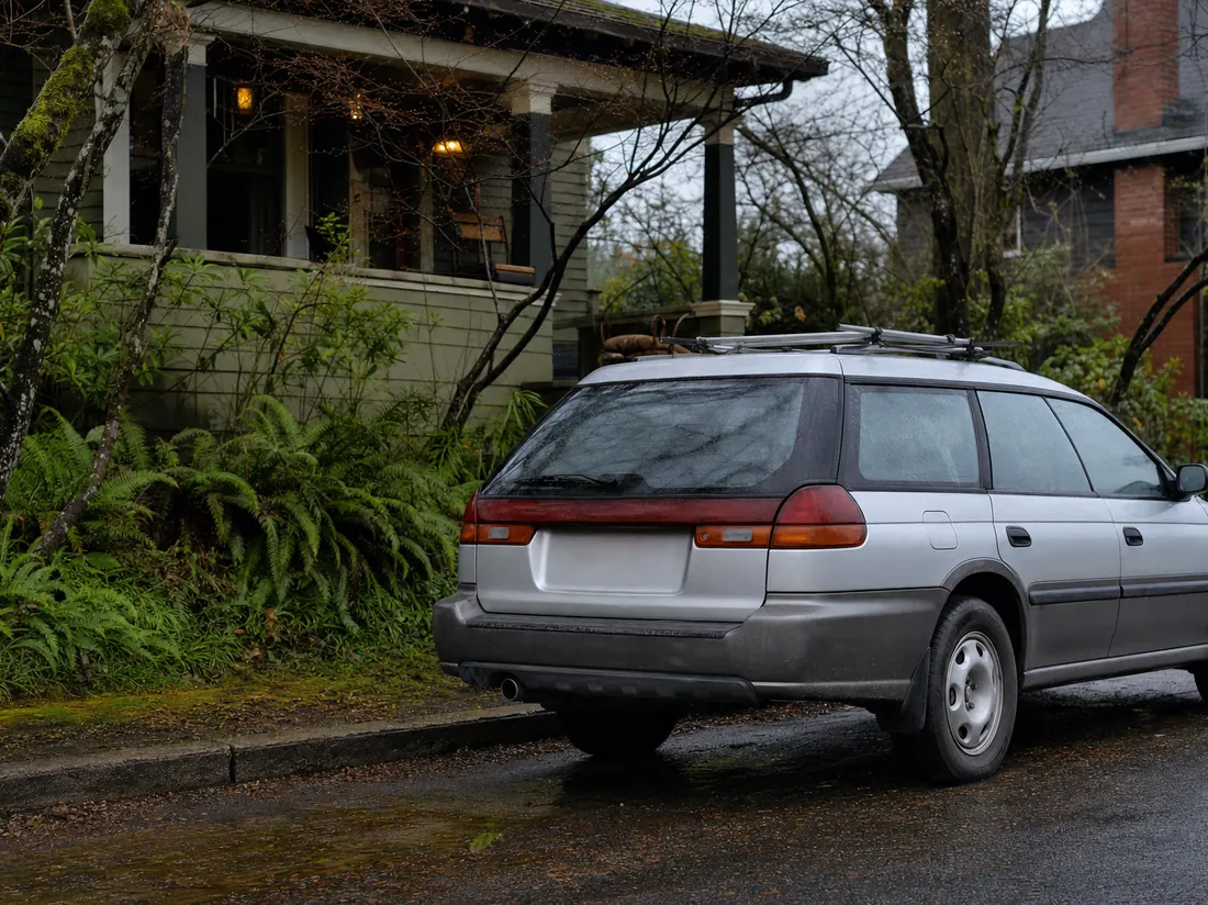 Older Subaru-style wagon with a faded bumper parked by a Portland craftsman with ferns and wet pavement reflections - selling a car in Portland