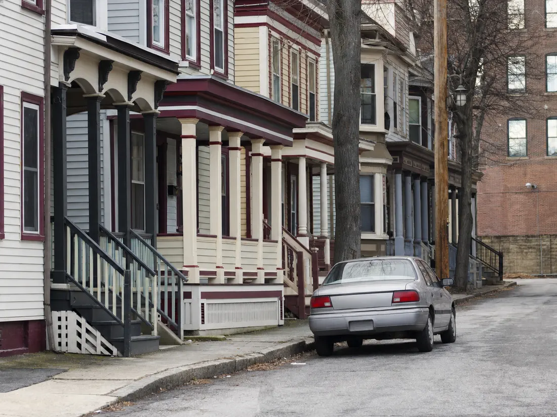 Older sedan on a dense street of mill-era duplexes in Pawtucket Rhode Island with painted porches and a brick mill wall beyond - selling a car in Pawtucket