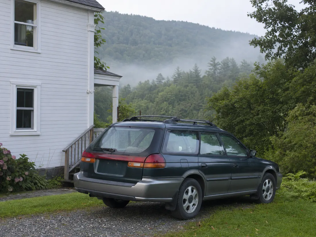 Older Subaru-style wagon by a white clapboard home in Montpelier Vermont with wooded green hills close behind - selling a car in Montpelier